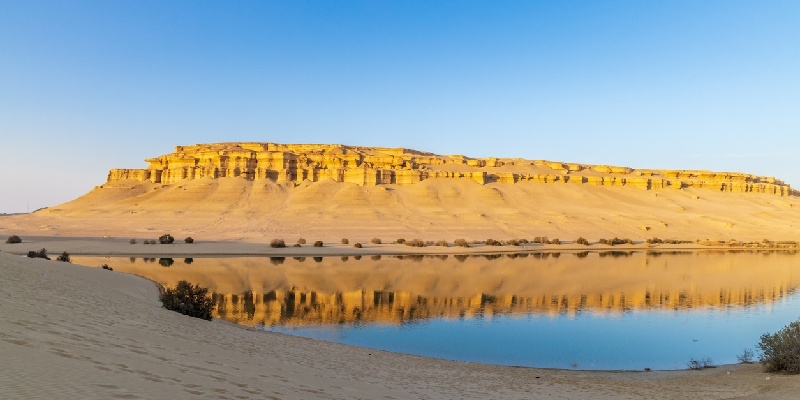 Fayum en Egipto Vista panorámica del paisaje desértico de Fayum, Egipto, con formaciones rocosas doradas reflejándose en un tranquilo lago en primer plano, bajo un cielo despejado.