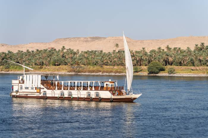 Traditional Dahabiya sailboat cruising along the Nile River with lush palm trees and desert hills in the background.